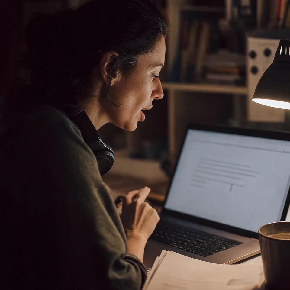 Close over-the-shoulder view of a woman speaking toward a laptop whose screen shows a text field with a sentence appearing and a cursor