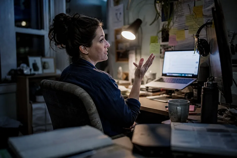 A woman leaning back from a laptop in a dim home office, speaking while an email draft fills the screen