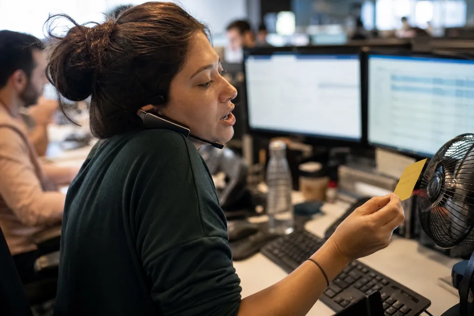 A support agent at a desk with a headset pushed back off one ear, speaking a reply while glancing at a sticky note